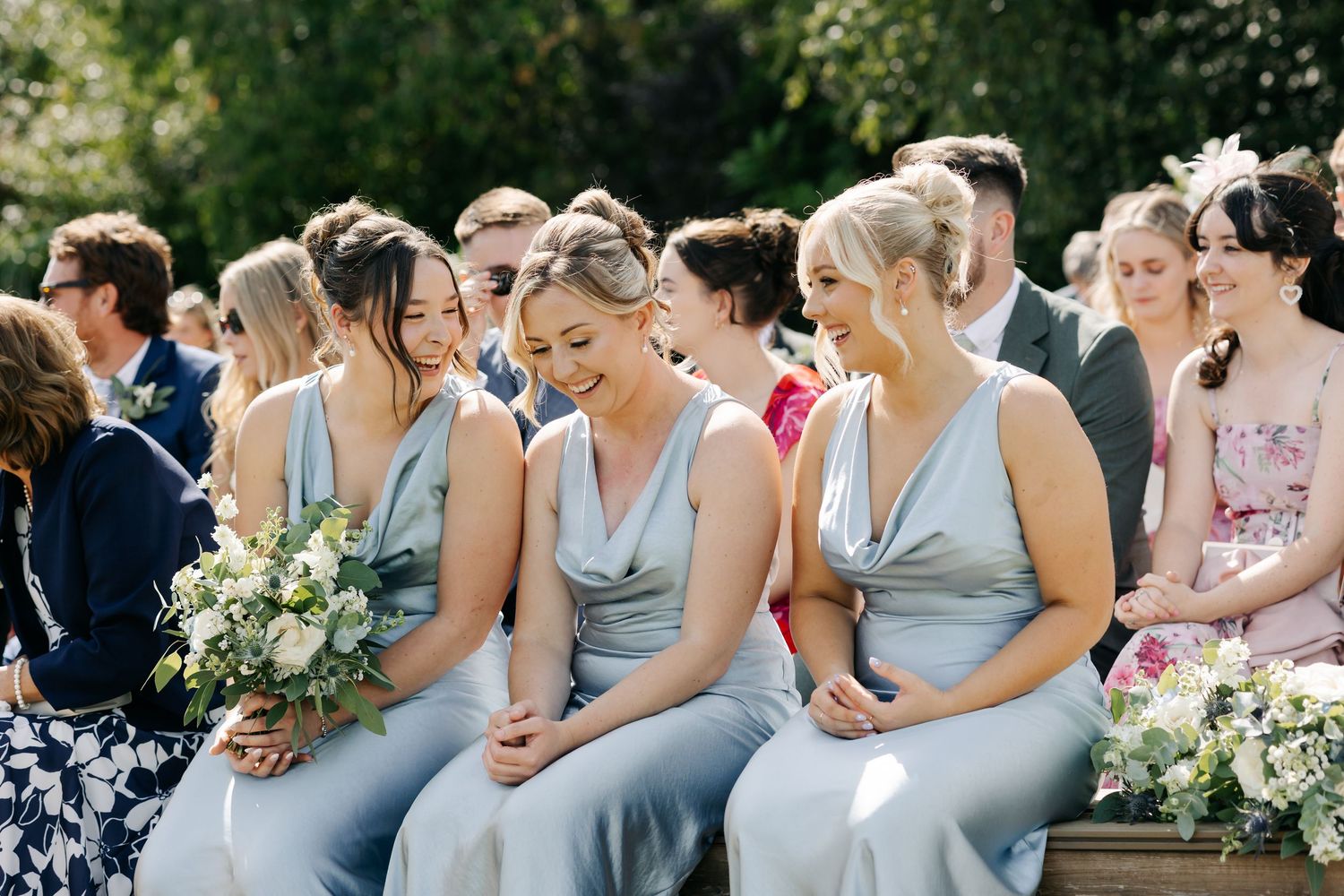 Wedding party members in light blue dresses sit together outdoors holding floral bouquets during a ceremony at Wickham Estate Hampshire