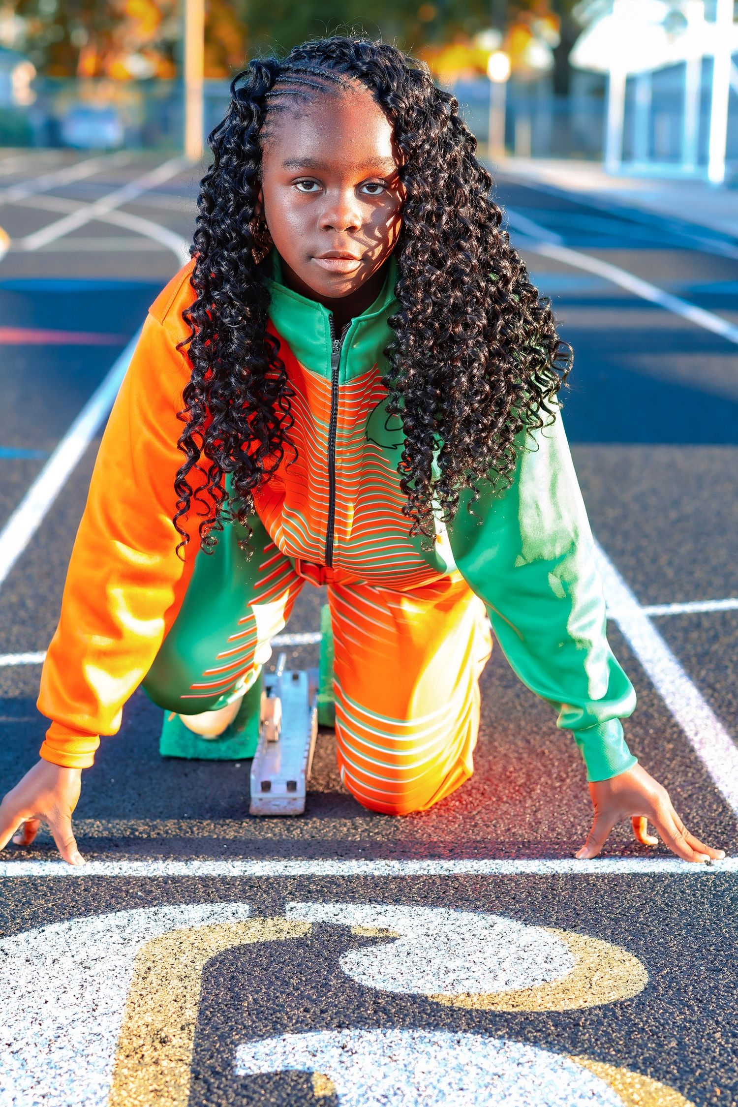 Runner in orange and green jacket crouches in starting position on asphalt track with painted white lines.