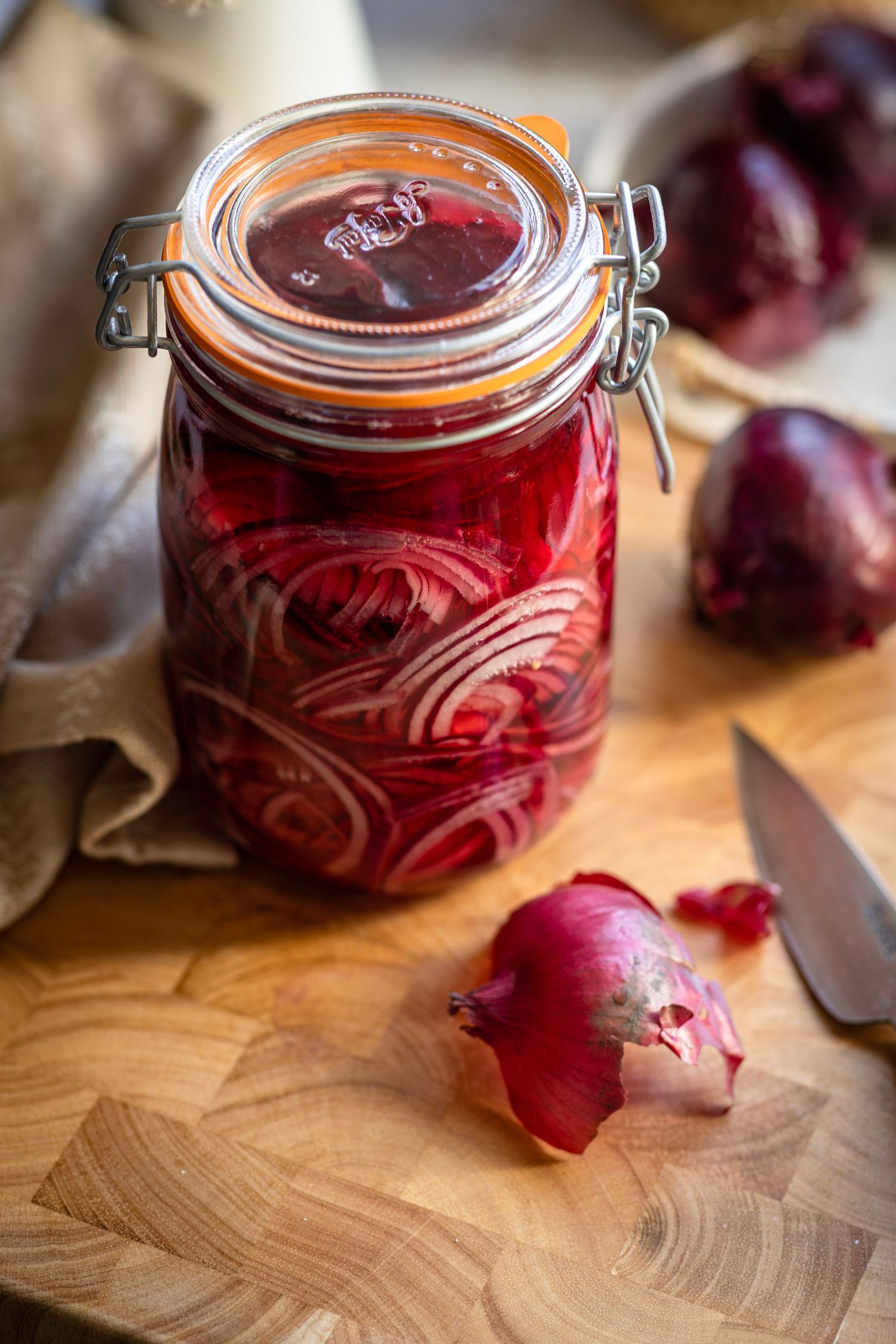 Freshly pickled red onions in glass jars on a wooden cutting board with raw onions nearby.