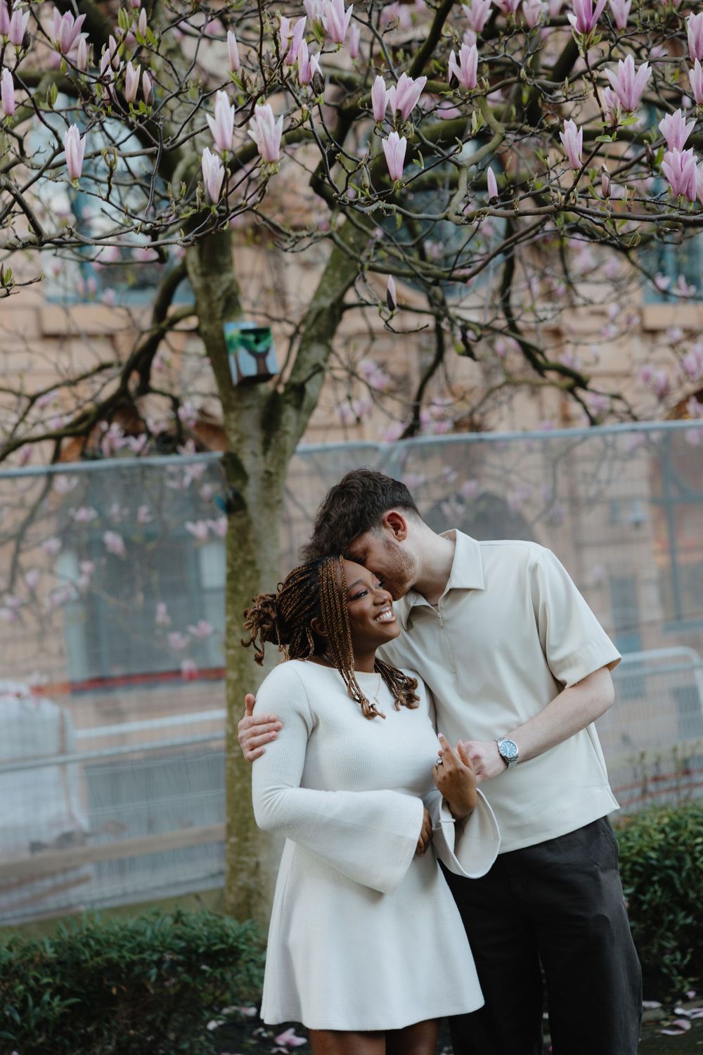 Two people in white outfits share a romantic moment under flowering magnolia branches.