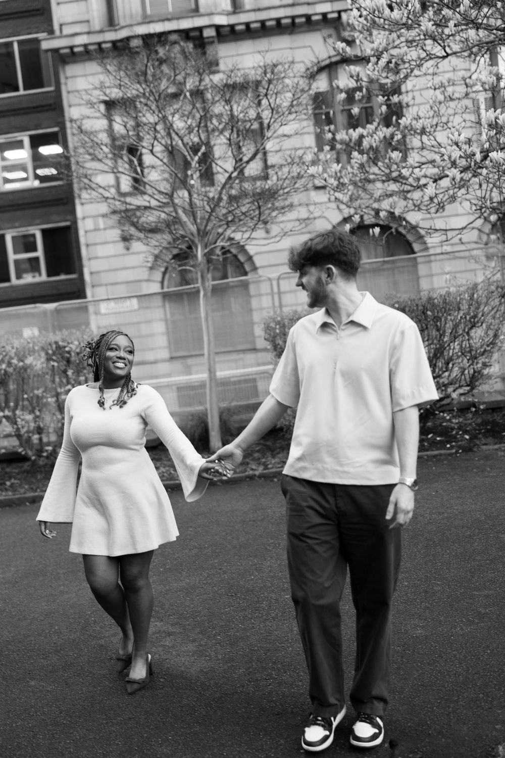 A joyful couple in matching white and black outfits dance playfully on a grassy lawn outside a building.