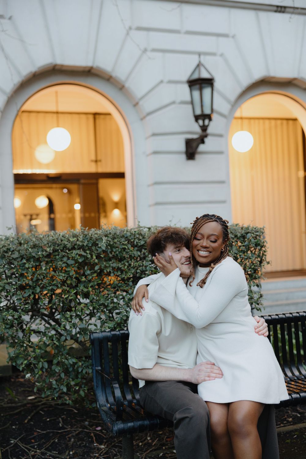 Two people in white sweaters embrace and laugh together on a bench near illuminated arches and greenery.