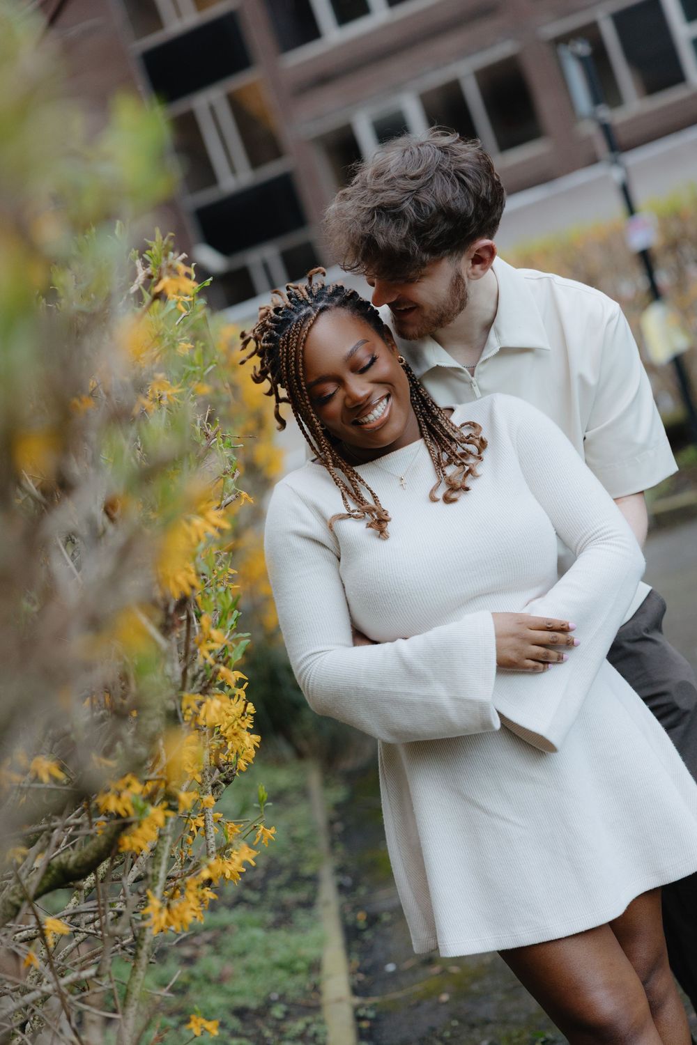 A couple in white sweaters embraces tenderly among yellow flowering branches in an outdoor setting.