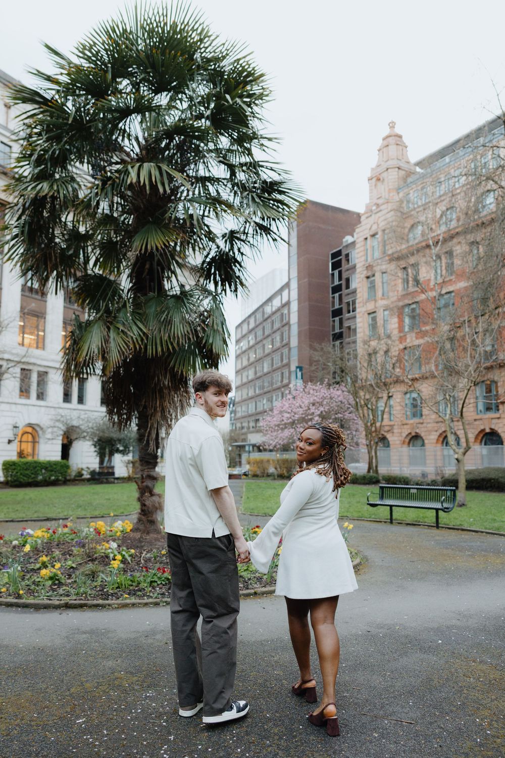 A couple shares a romantic moment in front of a historic building with palm trees and gardens in the background.