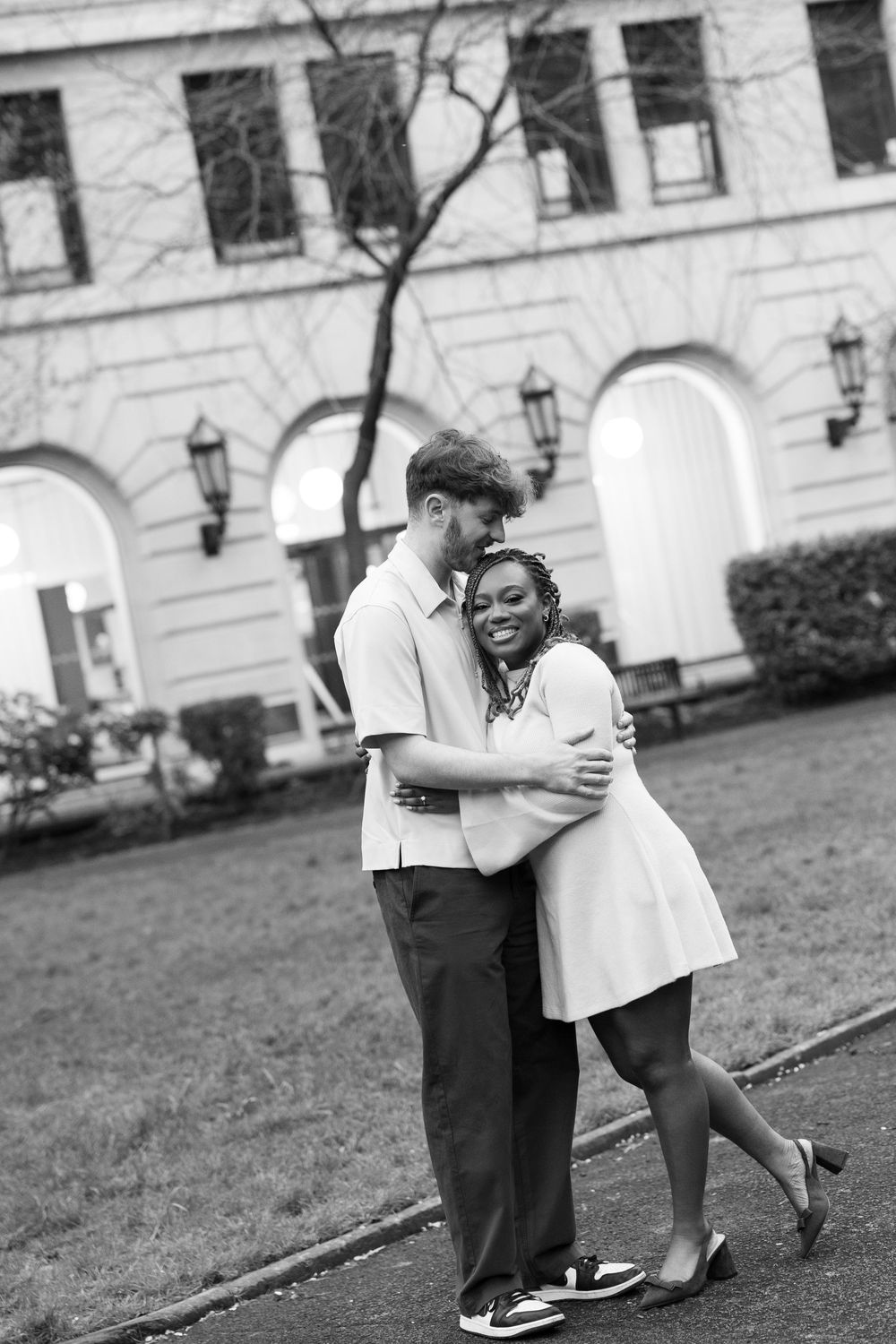 A romantic black and white photo of a couple embracing outdoors in front of a building with arched doorways and lanterns.