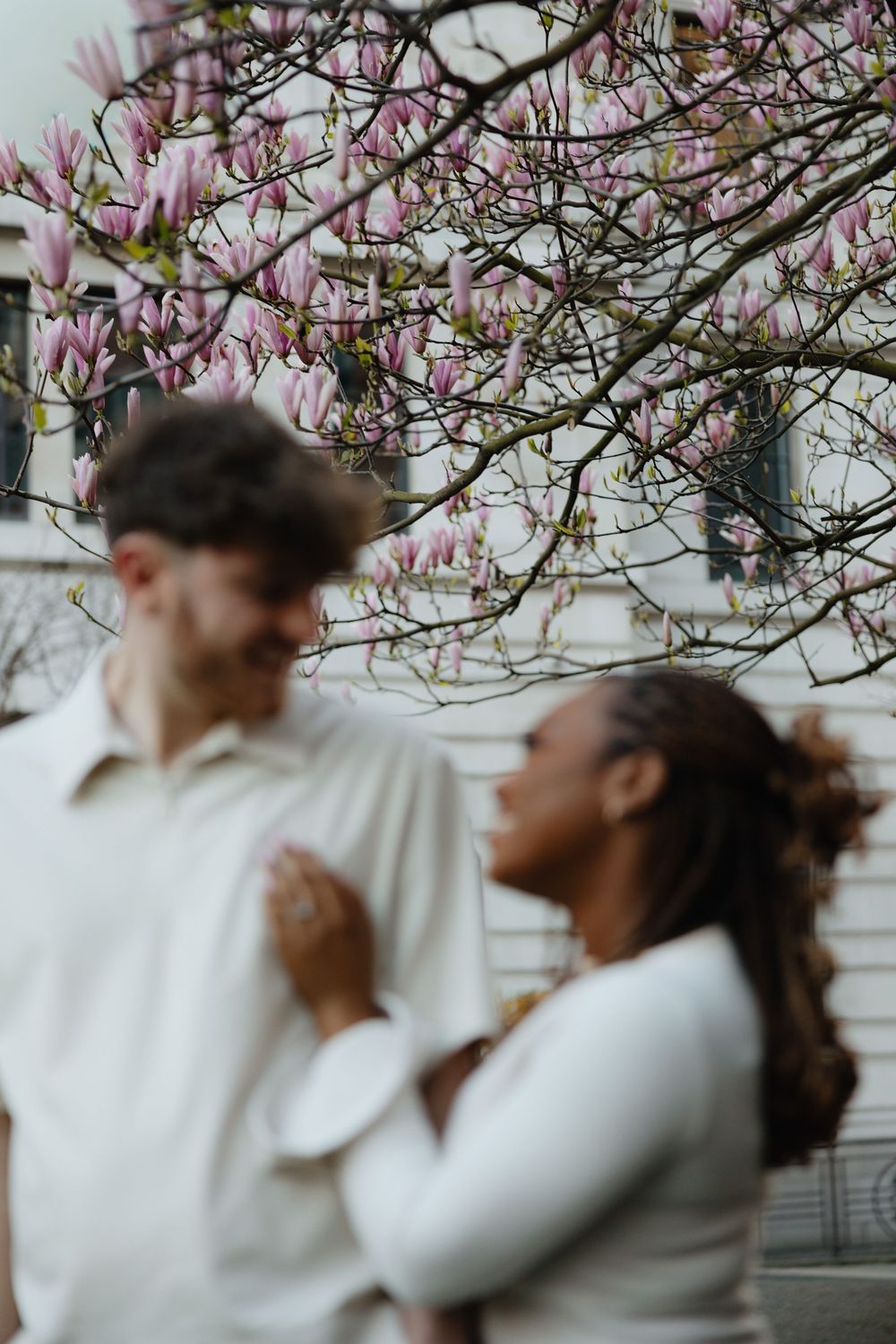 A couple poses beneath blooming pink magnolia blossoms in a soft, dreamy outdoor setting.