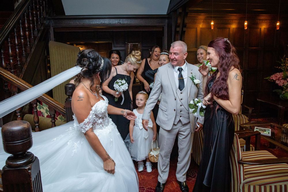 Bride greeted by groom and flower girl on wooden staircase surrounded by bridesmaids in navy dresses at wedding venue.