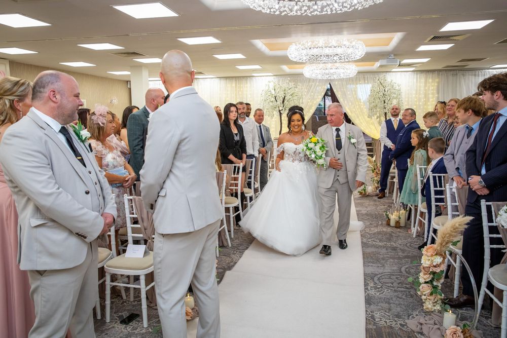 Bride walks down the aisle in white ball gown holding bouquet during elegant wedding ceremony with seated guests.