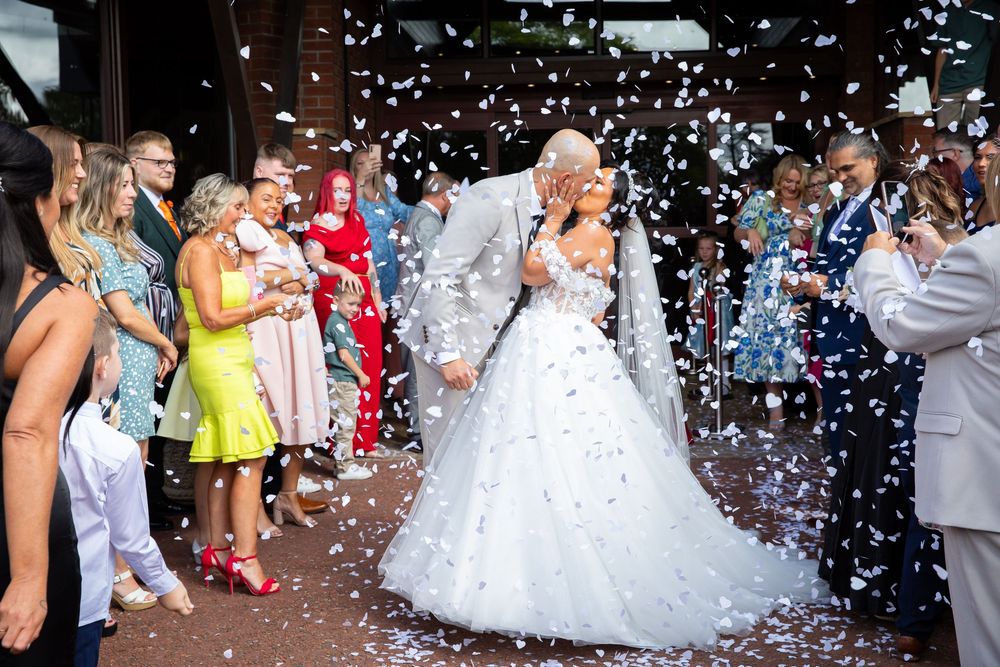 Newlyweds share a kiss surrounded by cheering guests throwing colorful confetti outside wedding venue.