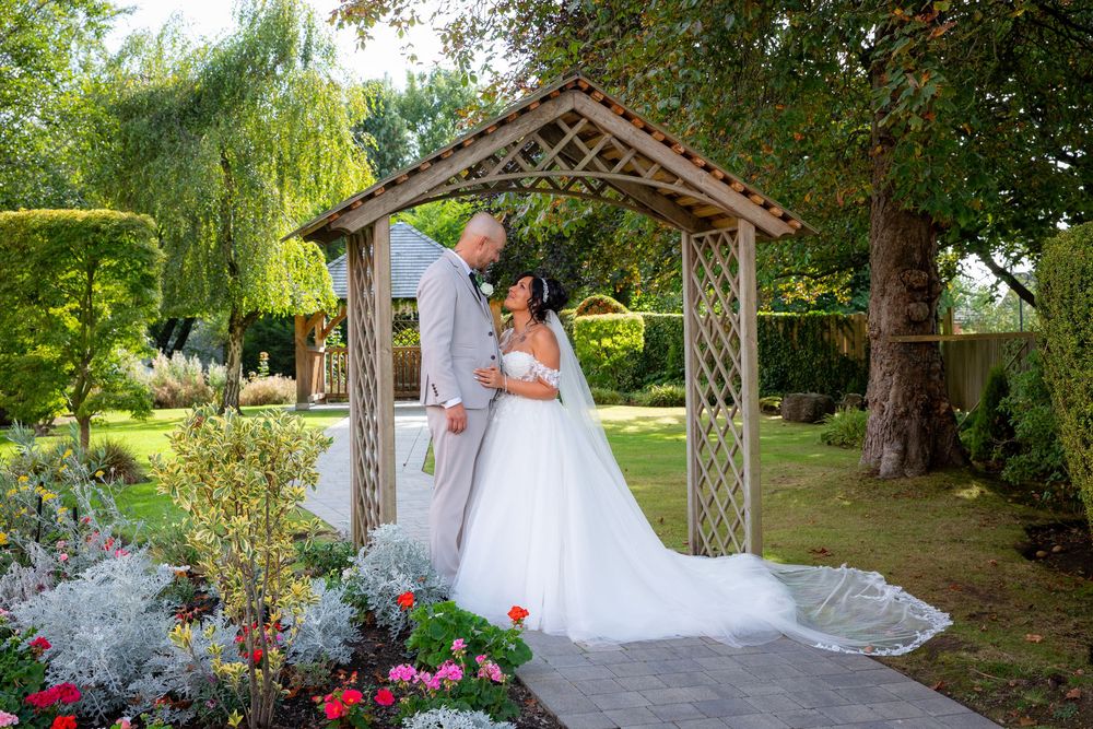 Bride and groom share romantic moment under wooden garden arbor surrounded by lush greenery and colorful flowers.