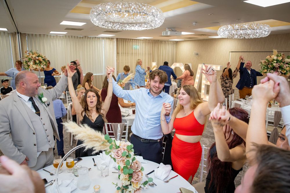 Wedding reception guests celebrate and raise glasses in elegant ballroom with crystal chandeliers and floral centerpieces.
