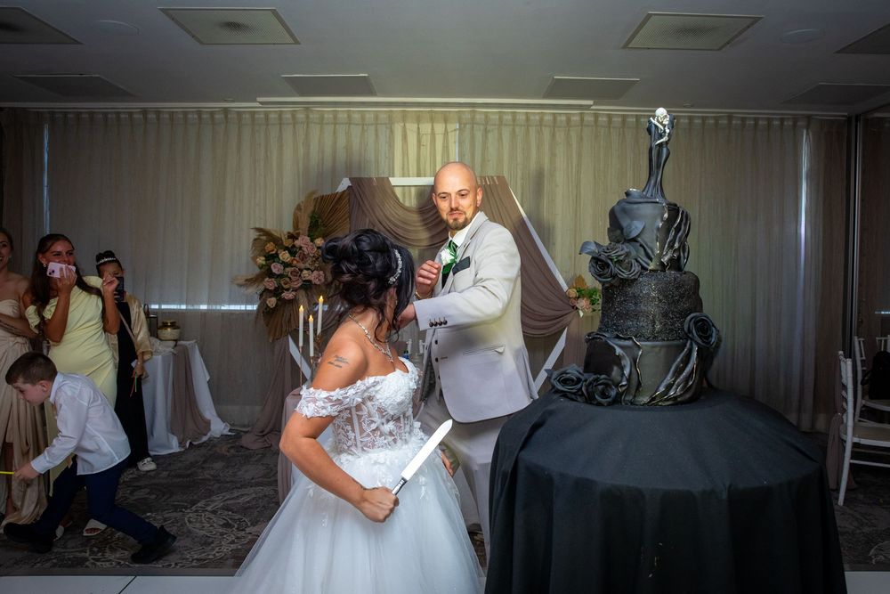 Bride and groom prepare to cut elaborate dark wedding cake during reception while guests photograph the moment.
