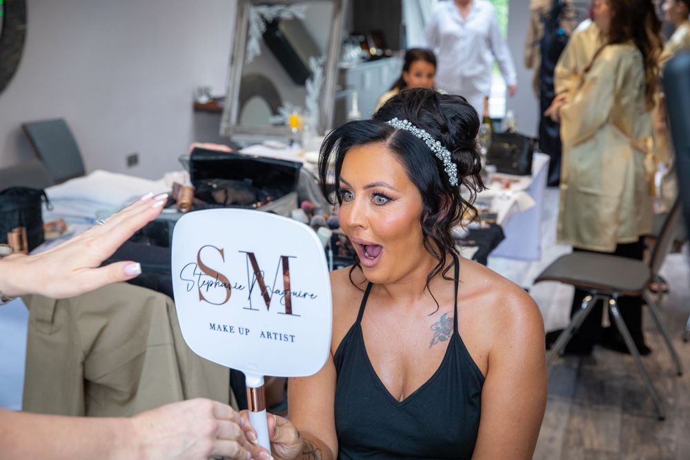 Bride reacts with surprise looking in branded makeup artist mirror during wedding morning preparations.