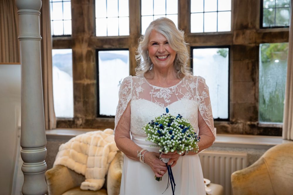 Radiant mature bride smiles holding a delicate blue and white bouquet in a beautifully lit venue.