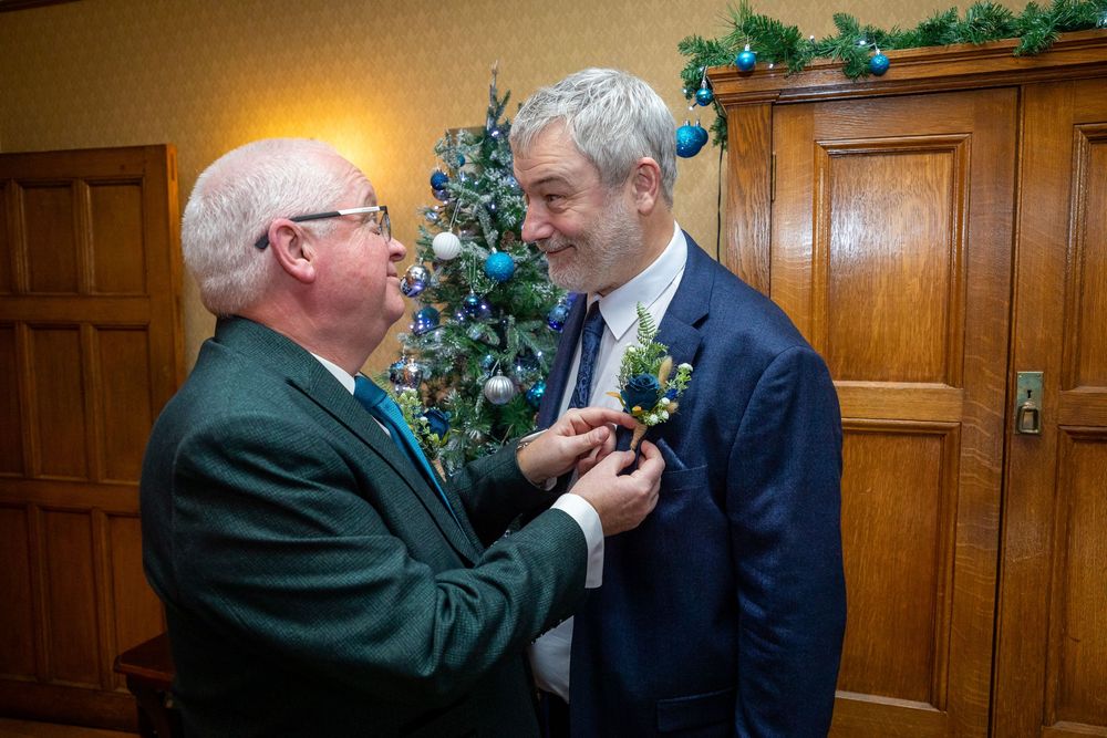 Man pinning a boutonniere on groom's lapel near a Christmas tree at a winter wedding.