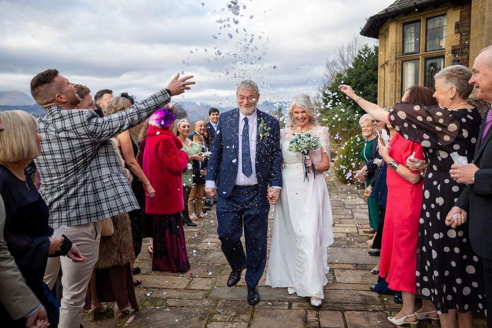 Newlyweds walking through confetti shower outside a stone venue surrounded by cheering wedding guests.