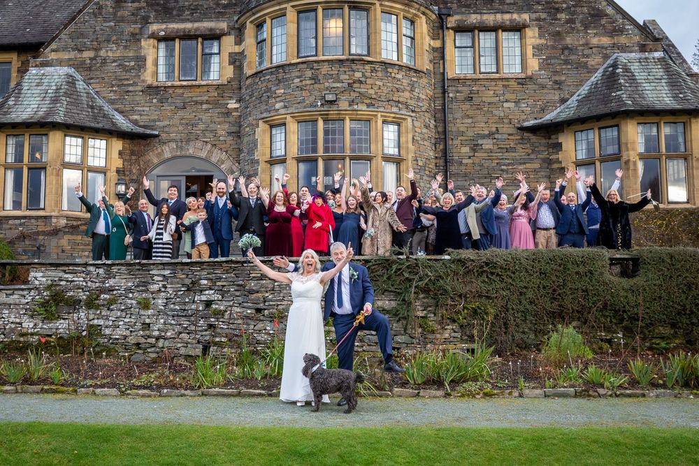 Bride and groom pose with dog and cheering wedding guests in front of a grand stone manor house.