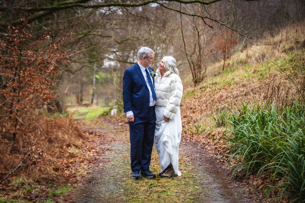 Couple share a romantic moment on a woodland path, bride in white gown and groom in navy suit.