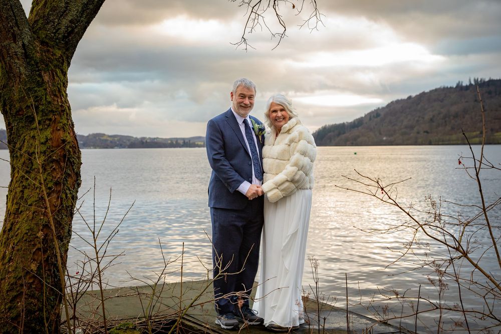 Elegant bride and groom pose hand in hand by a serene lakeside under a dramatic cloudy sky.