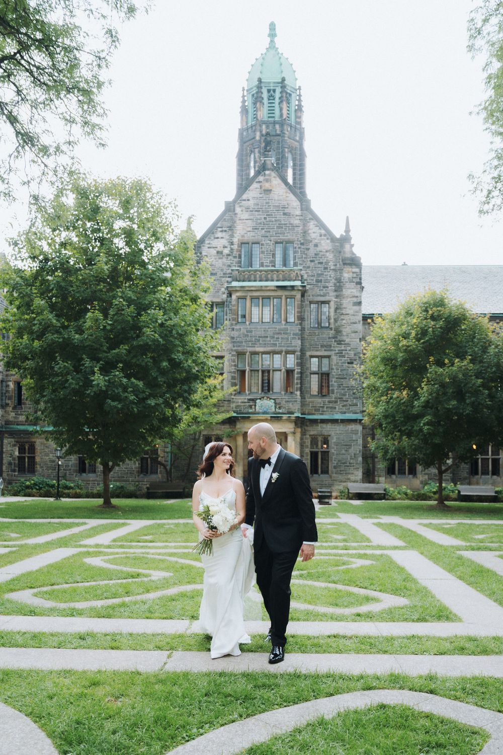 Bride and groom walk hand-in-hand on manicured grounds before a grand Gothic stone building on their wedding day.