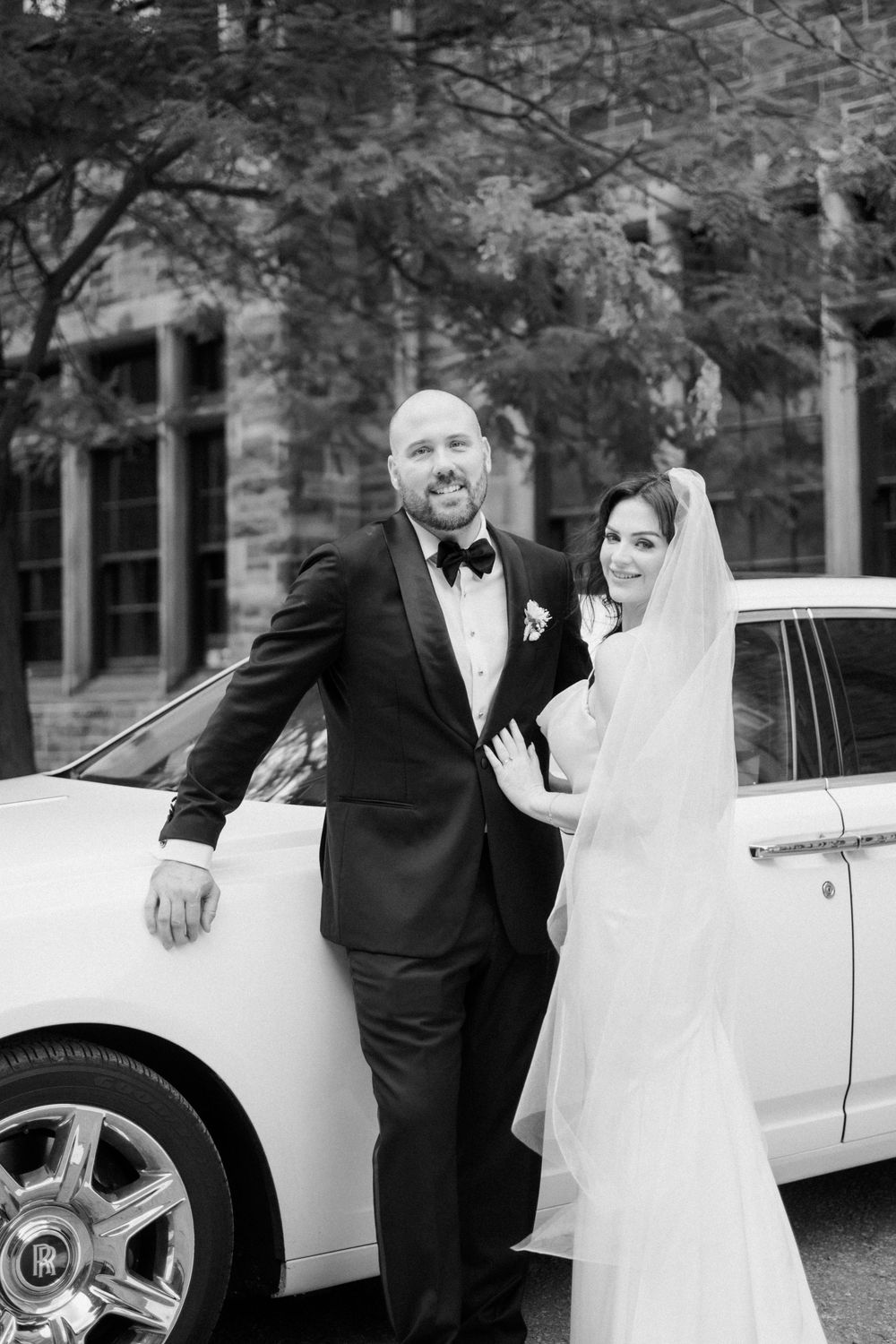 Black and white wedding photo of bride and groom posing elegantly beside a white luxury car outside a stone building.