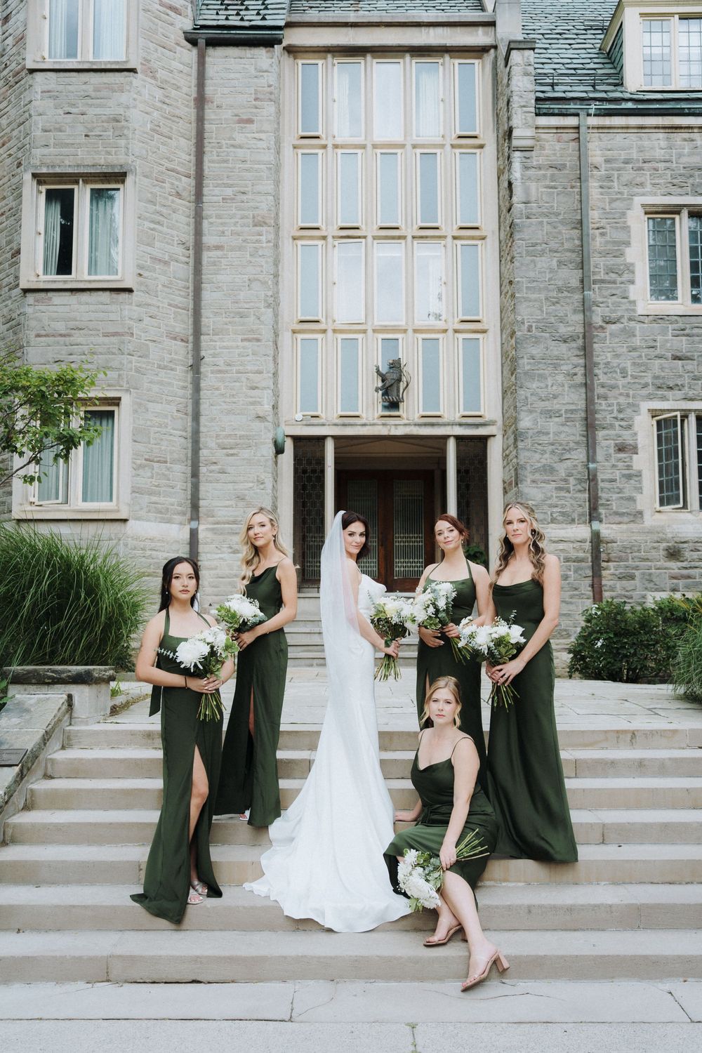 Bride in white gown with bridesmaids in dark green dresses holding bouquets on stone steps of elegant building.