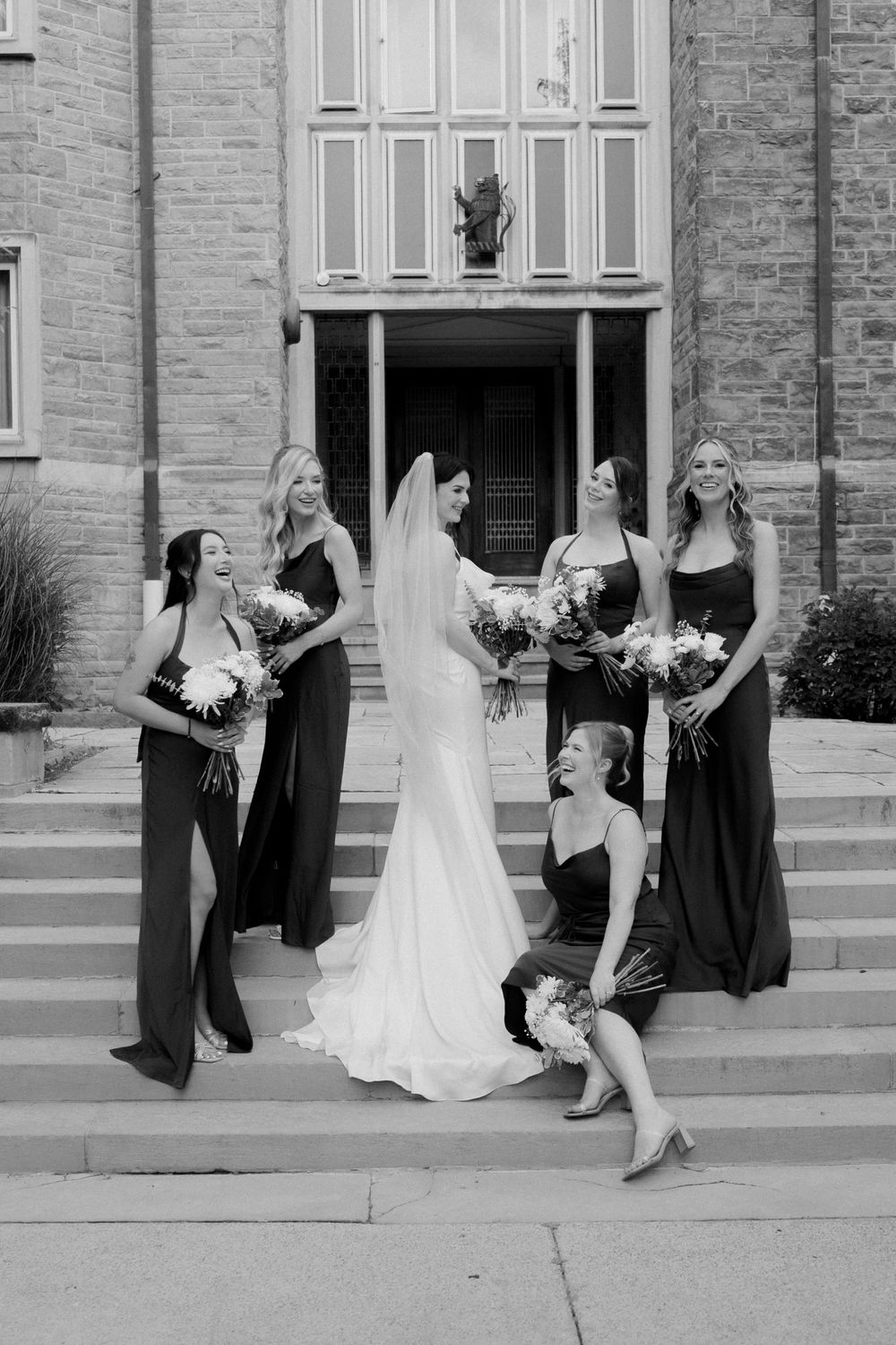 Black and white photo of a bride in a white gown and veil with four bridesmaids in dark dresses on church steps.