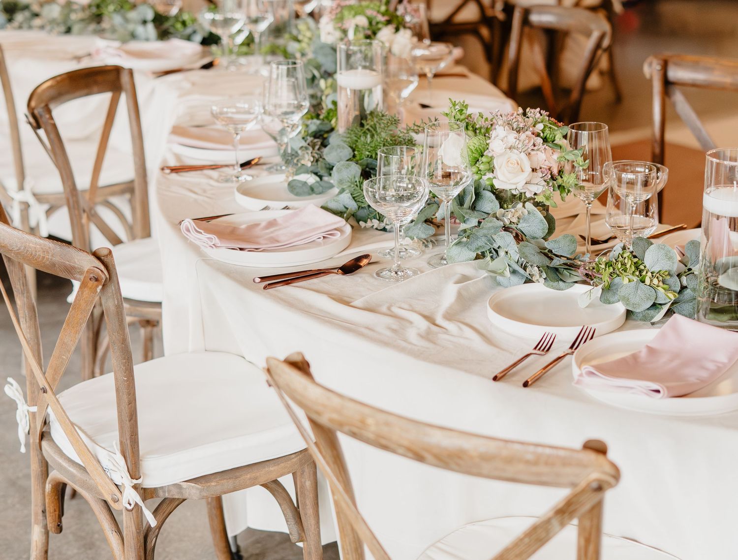 Elegant wedding reception table with blush pink napkins, eucalyptus garland, floral centerpieces, and rustic wooden cross-back chairs.