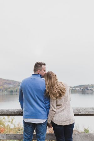 A couple in blue and beige sweaters look out over a lake from a viewpoint on an overcast day.