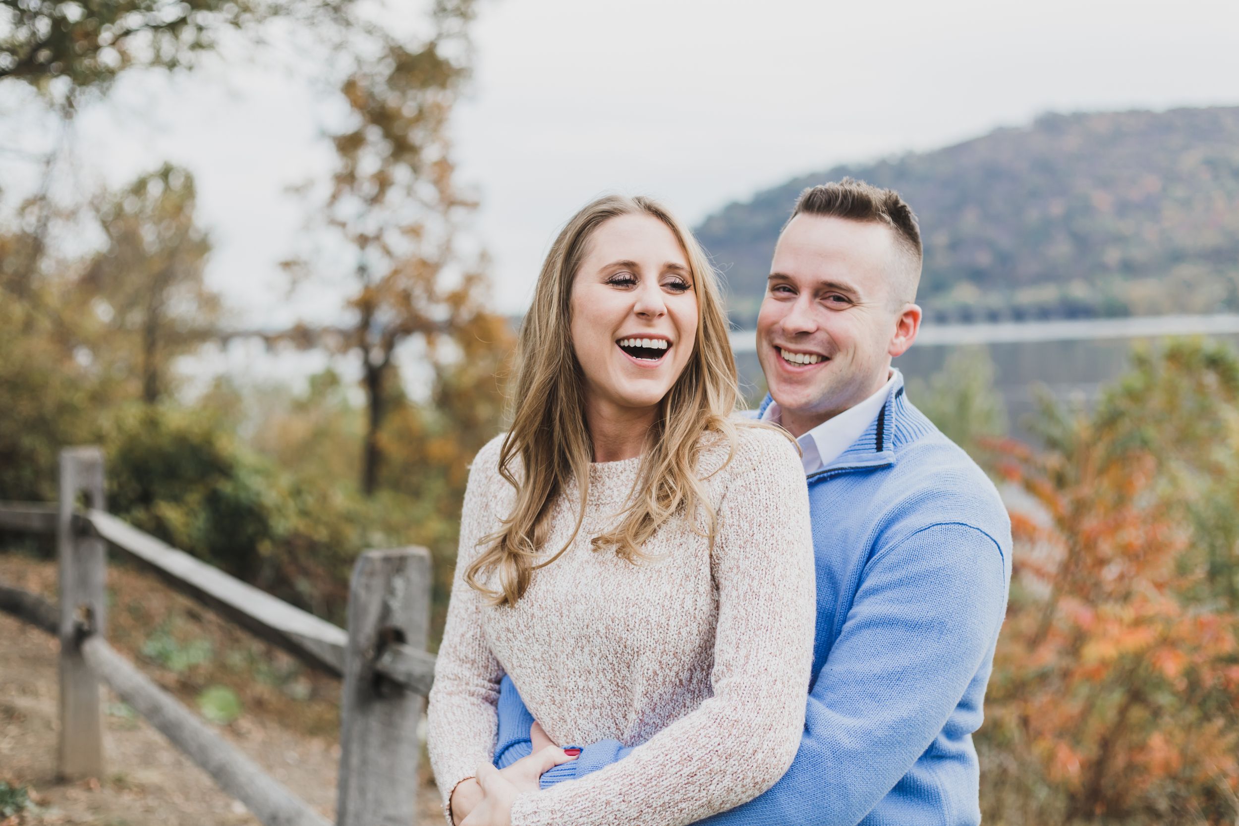 A couple embraces outdoors during an autumn engagement photoshoot with mountains and fall foliage in the background.