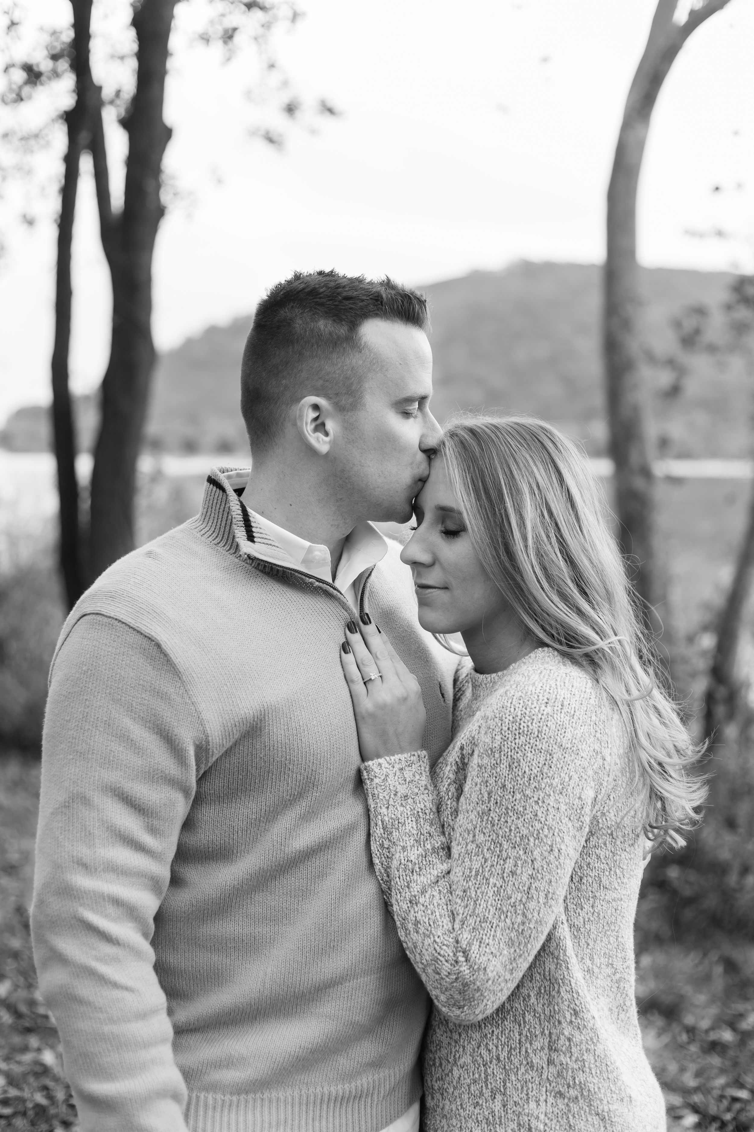 A romantic black and white portrait of a couple embracing outdoors in a wooded setting.