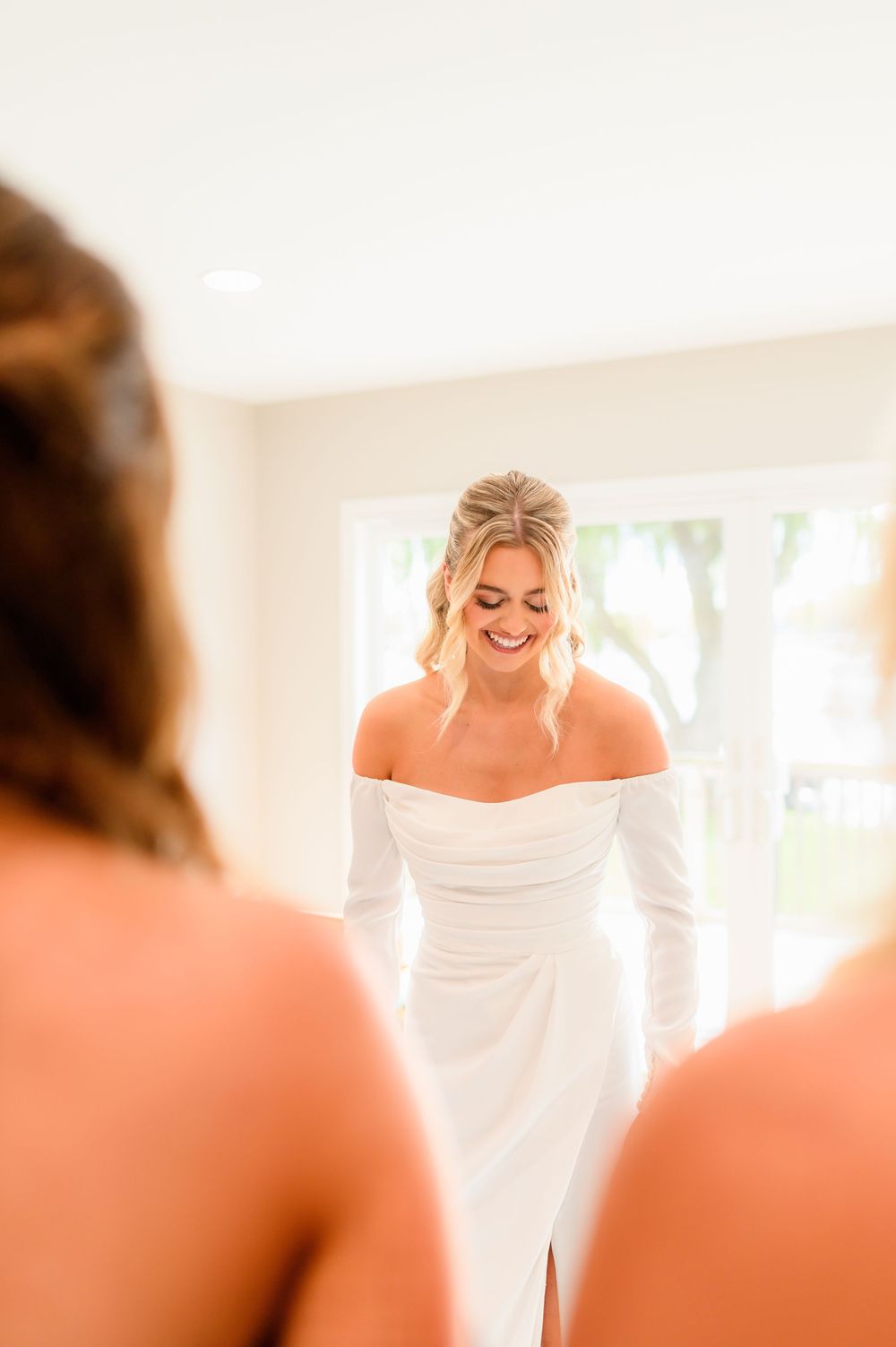Smiling bride in off-shoulder white gown laughing with bridesmaids in bright room.