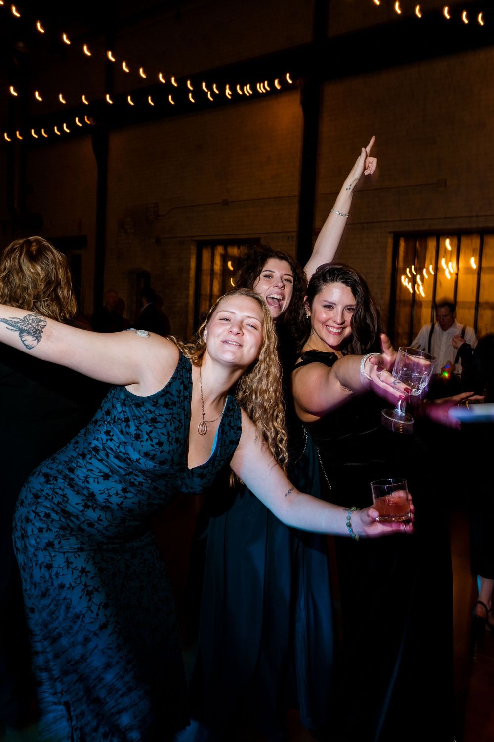 Three women in elegant black dresses celebrate at a wedding reception, dancing and raising drinks under string lights.