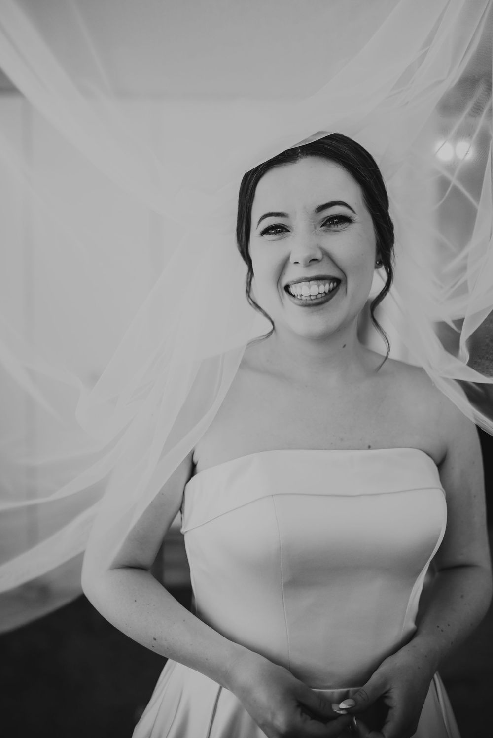 Black and white photo of a smiling bride in a strapless wedding gown with veil flowing behind her.