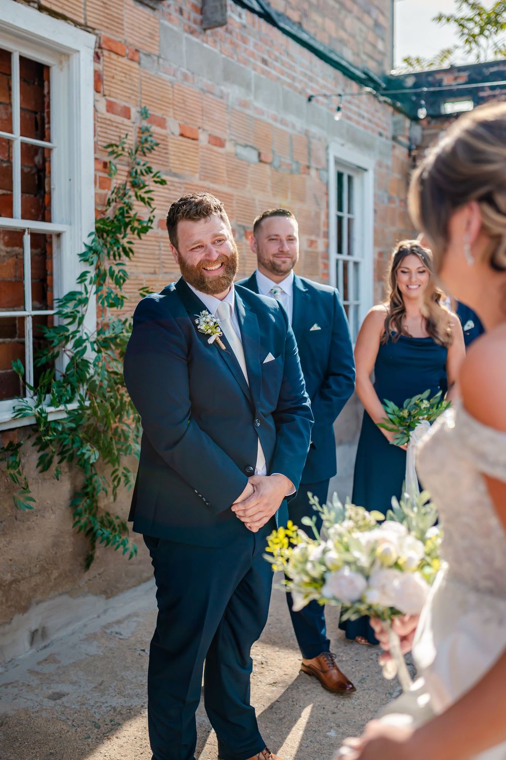 Groom smiling joyfully as bride approaches during outdoor wedding ceremony, with wedding party in navy blue attire.