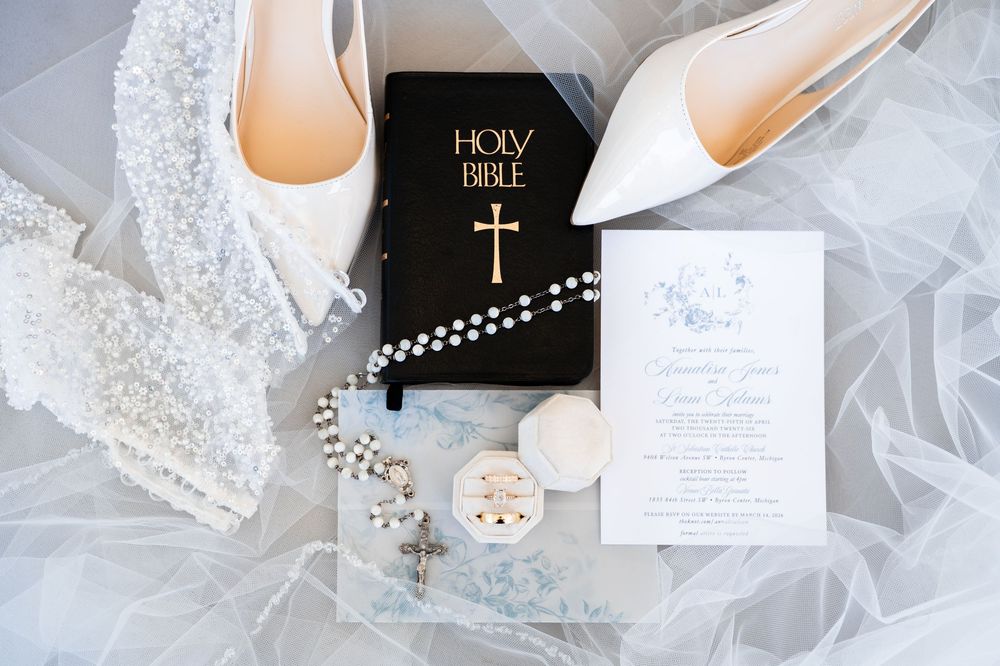 Elegant wedding flat lay featuring white heels, Holy Bible, rosary beads, lace veil, and floral invitation on sheer fabric.