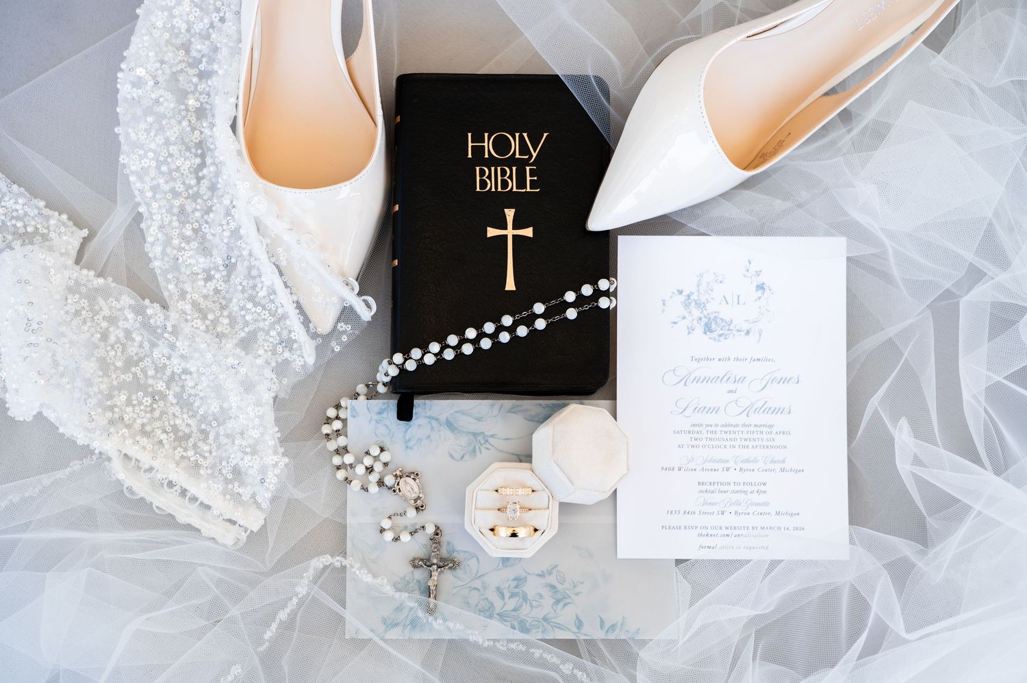 Elegant wedding flat lay featuring white heels, Holy Bible, rosary beads, lace veil, and floral invitation on sheer fabric.