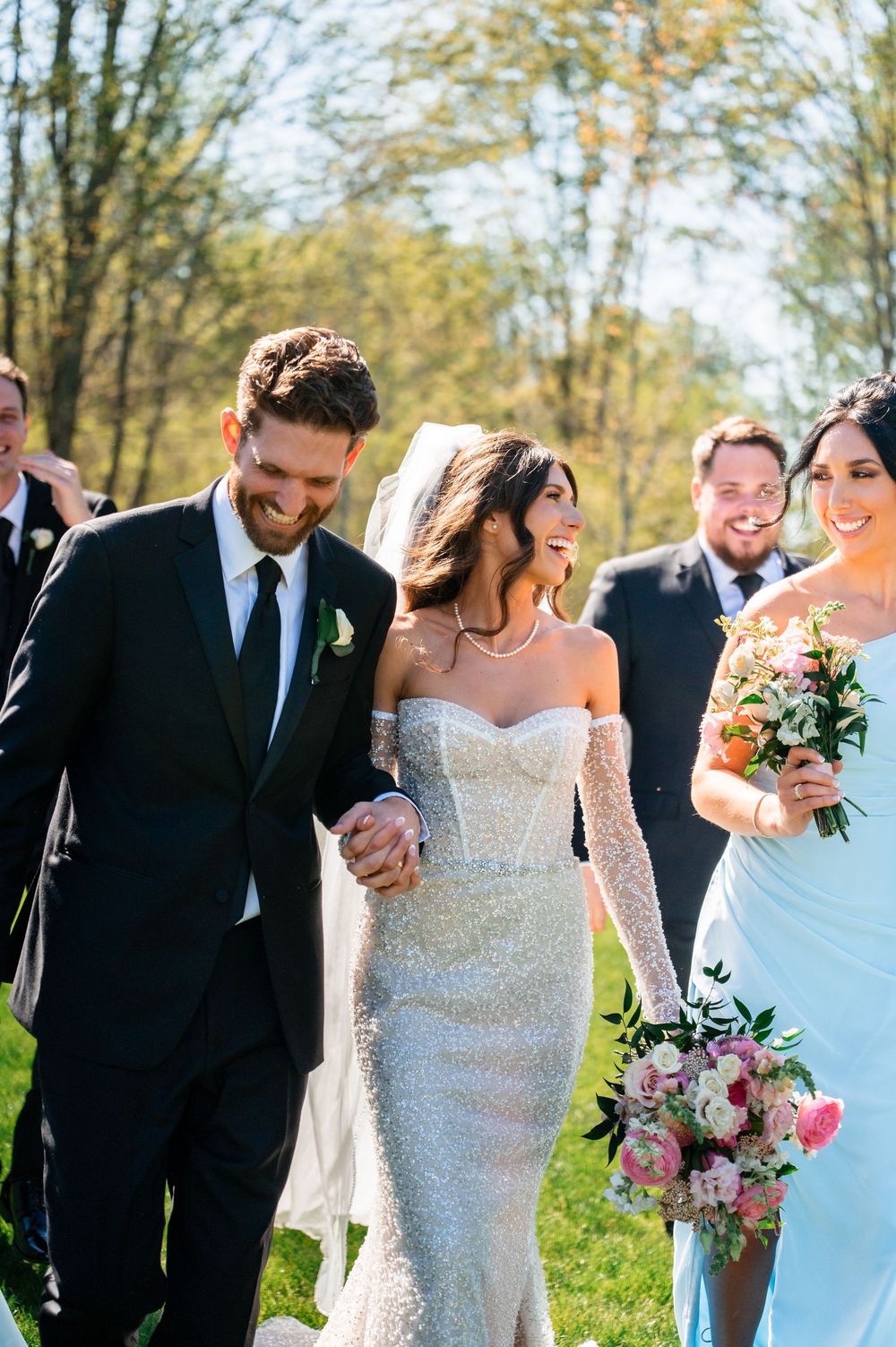 Joyful newlywed couple holding hands walking with wedding party outdoors on sunny day, bride in beaded off-shoulder gown.