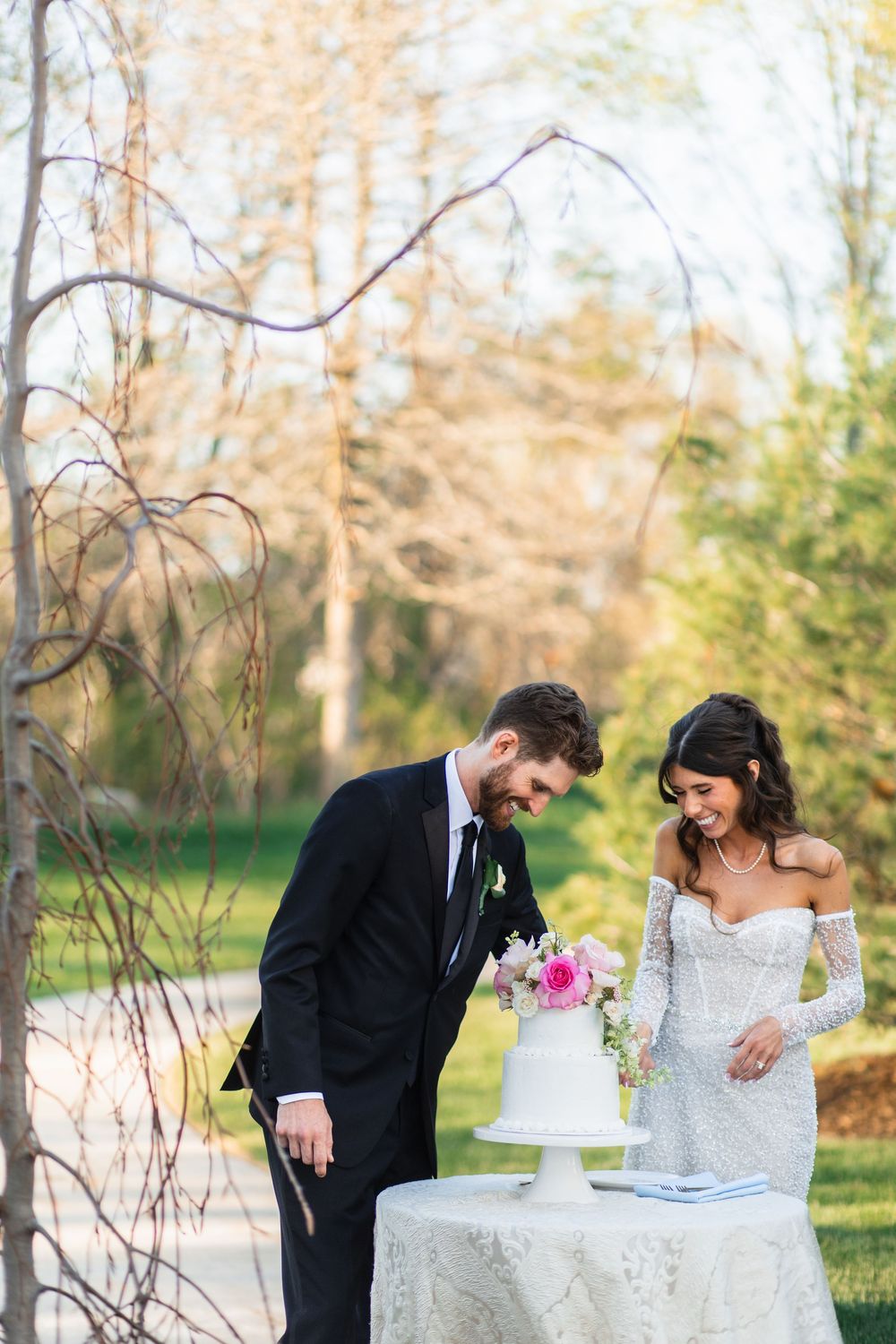 Bride and groom smiling while cutting white wedding cake outdoors surrounded by trees in natural garden setting.