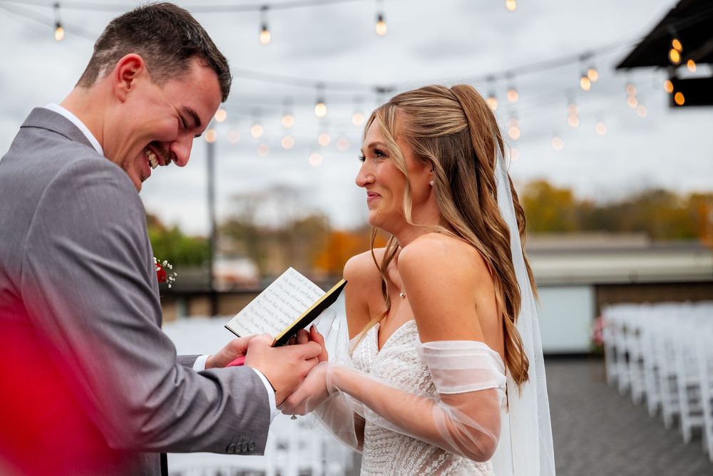Bride and groom laughing joyfully during outdoor wedding vows ceremony with string lights in background.