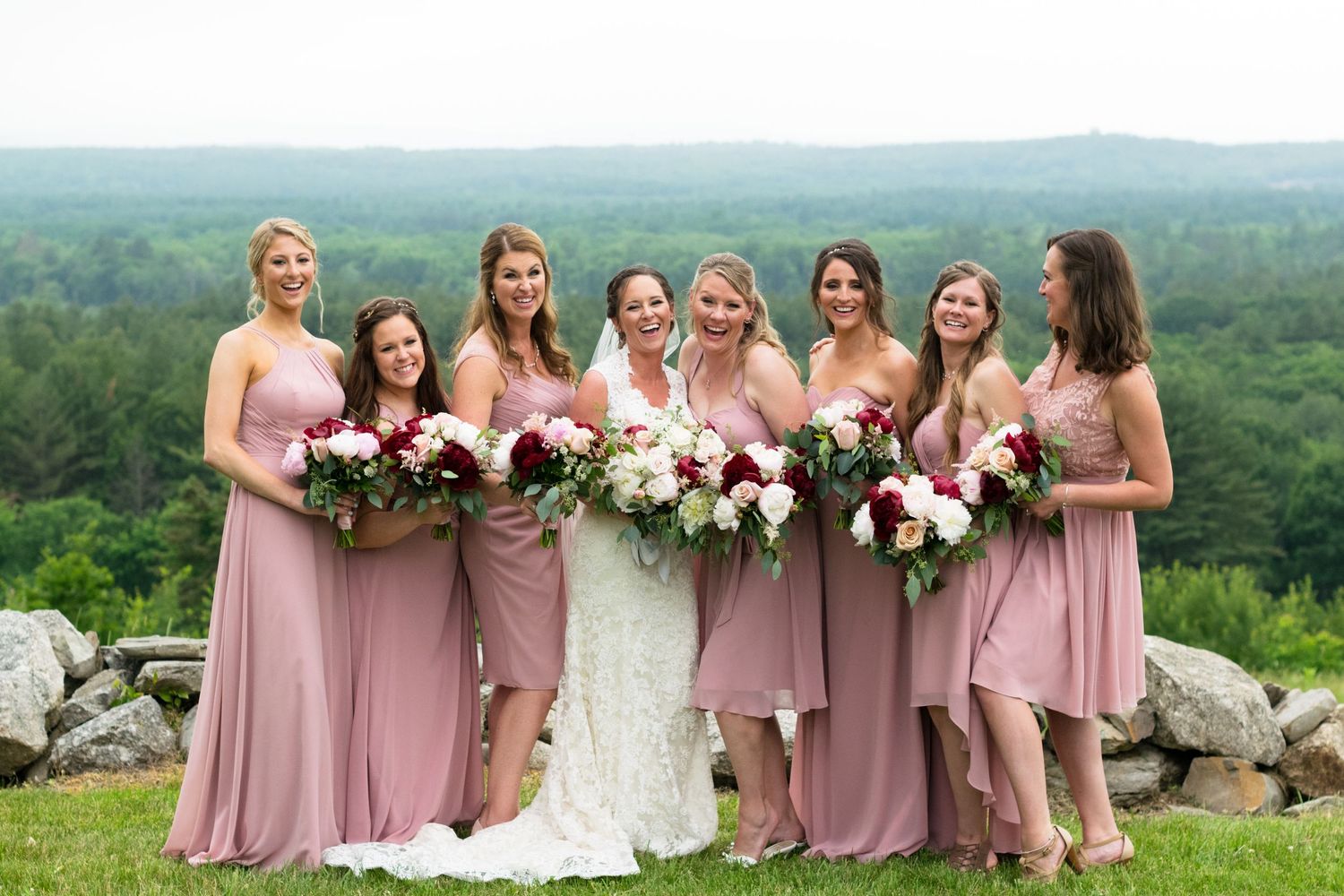 Bride in white lace gown poses with six bridesmaids in dusty rose dresses holding red and white bouquets outdoors.