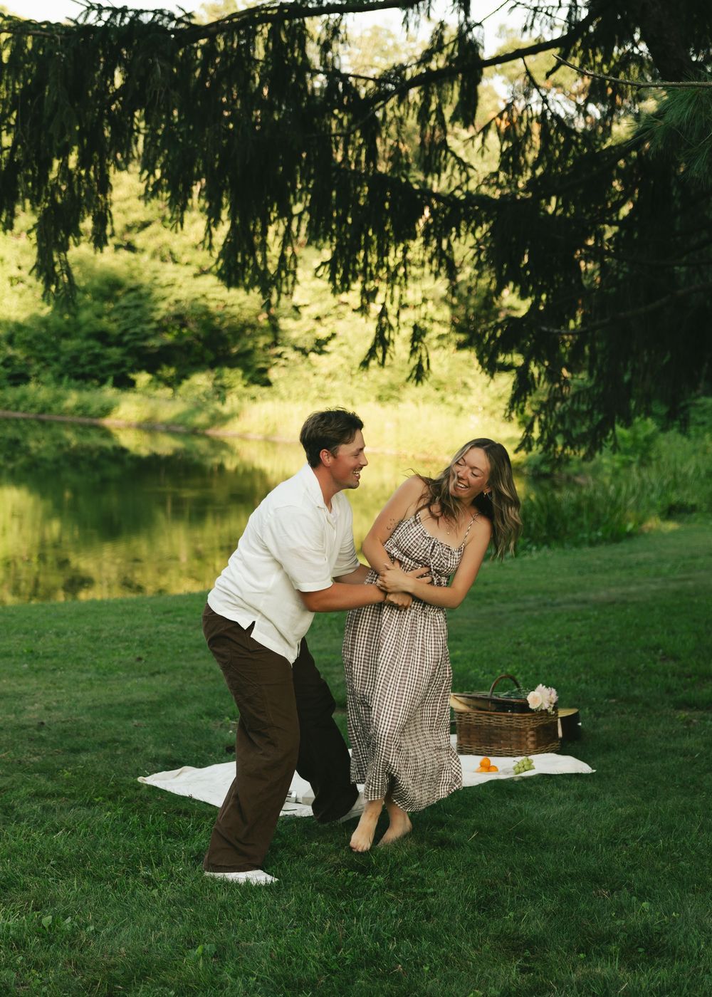 A joyful couple dancing and laughing together on a blanket by a serene pond during a romantic outdoor picnic.