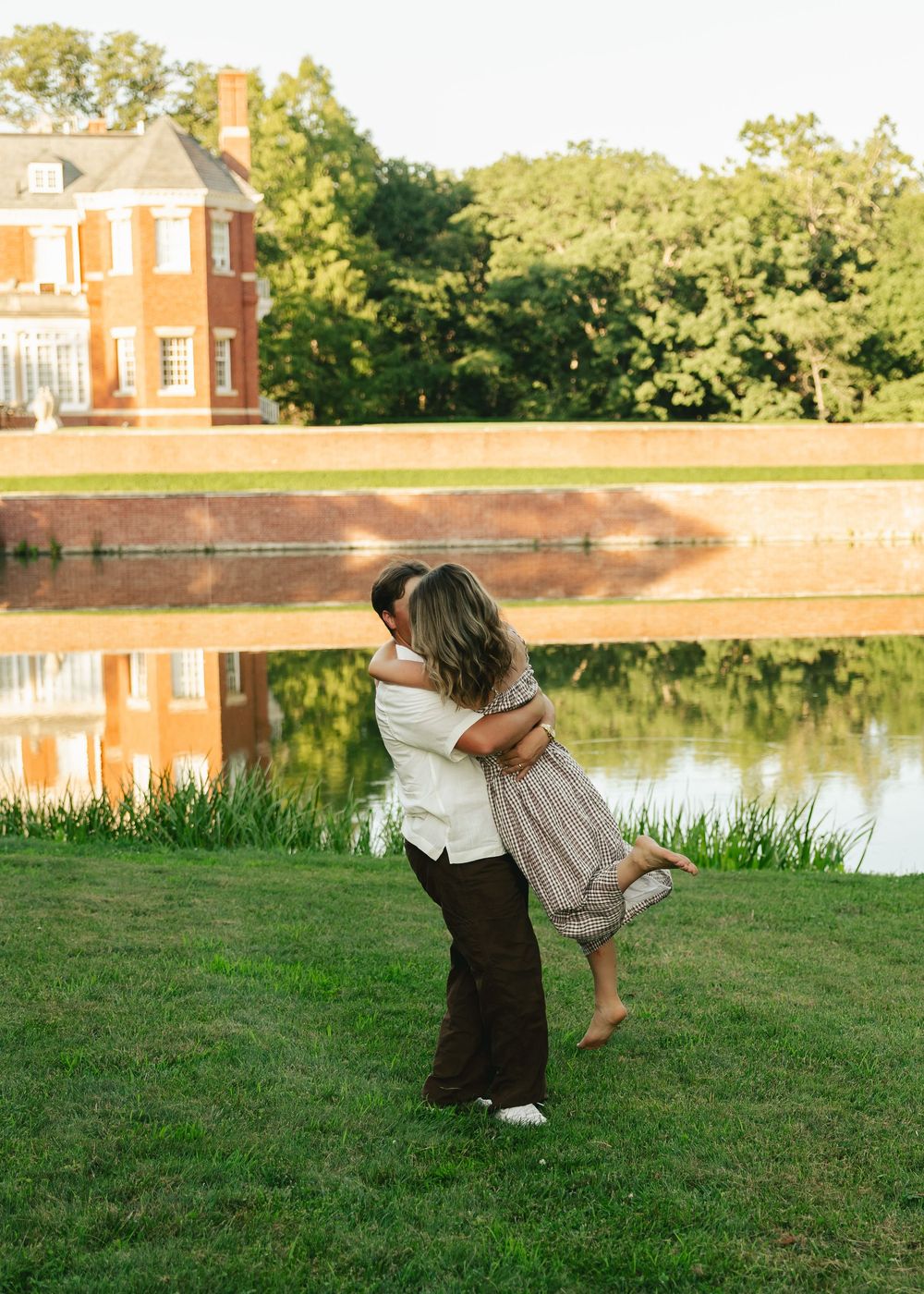 Couple embracing by a reflective pond with a brick mansion and lush trees in the golden hour light.