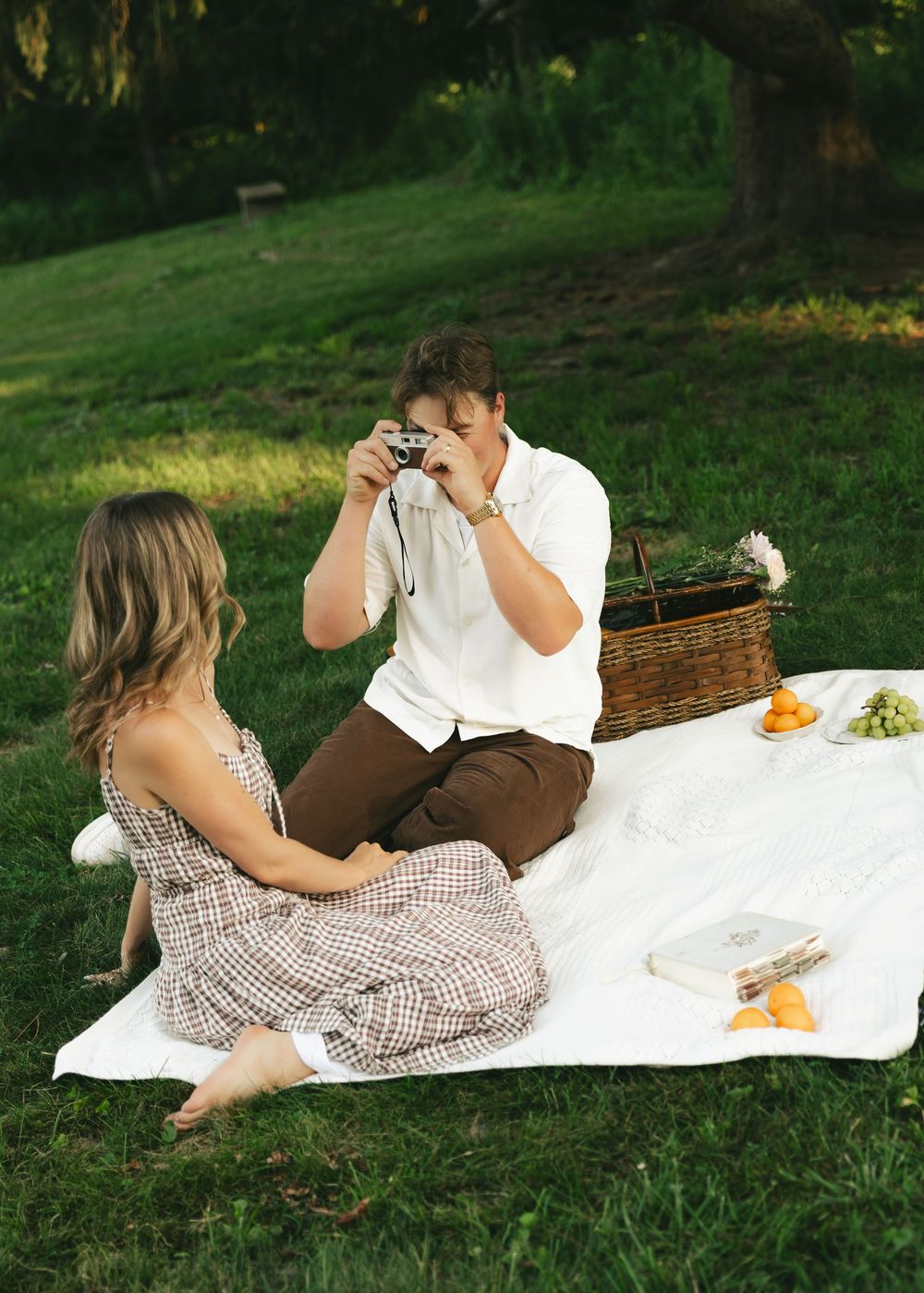 A couple enjoys a romantic outdoor picnic on a white blanket, with a man photographing a woman in a checkered dress.