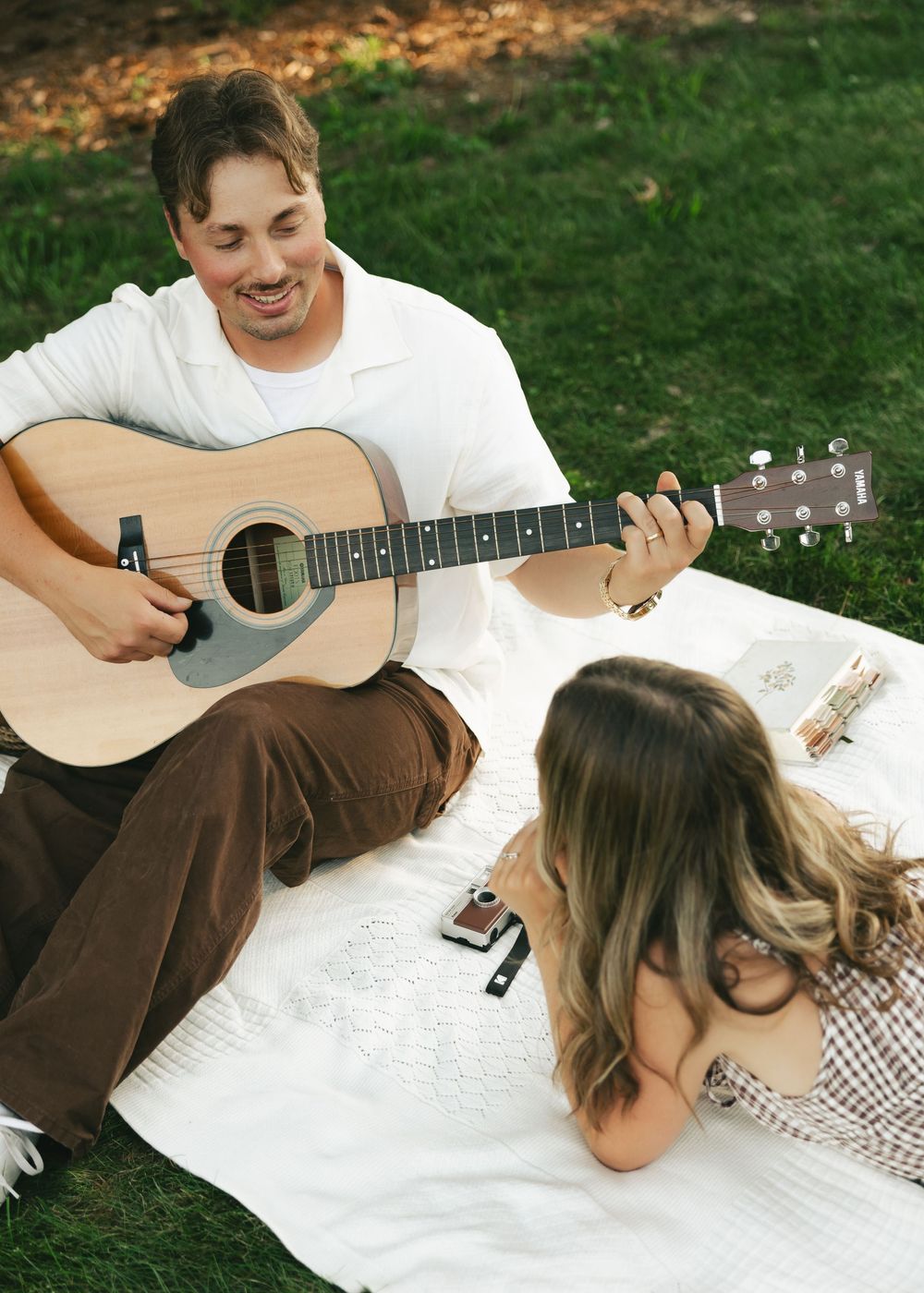Man playing acoustic guitar for woman on picnic blanket in park, romantic outdoor music date.