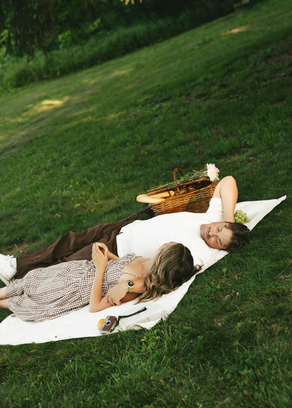 Couple lying on white blanket in green park during romantic picnic with wicker basket.