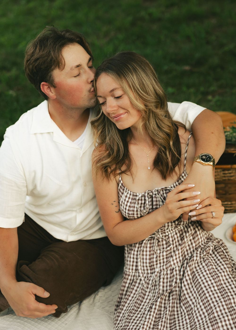 Couple sharing a romantic kiss during an outdoor picnic photoshoot, woman in gingham dress smiling.