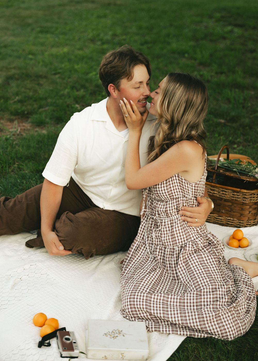 Couple sharing a romantic kiss during a picnic on a white blanket in a lush green park with a wicker basket.