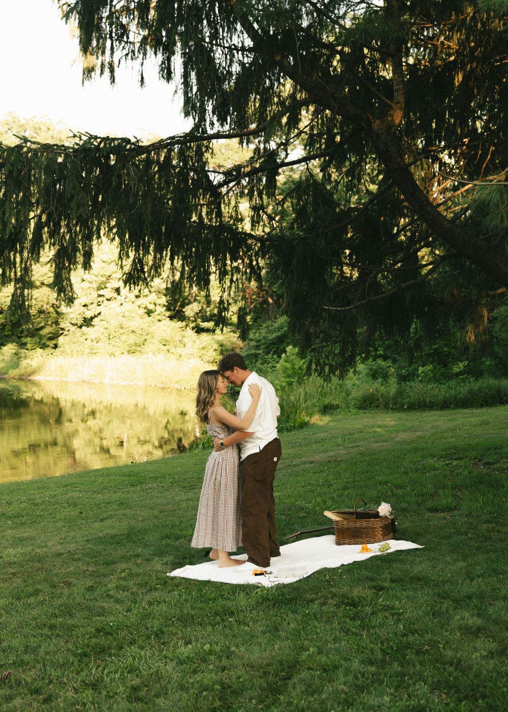 Couple sharing a romantic kiss on a white blanket by a serene pond under a weeping tree during golden hour.
