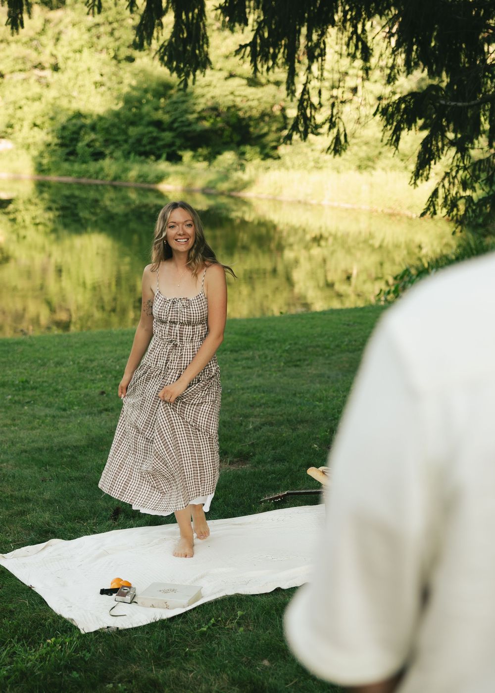 A smiling woman in a plaid sundress stands barefoot on grass near a serene pond during a summer picnic.